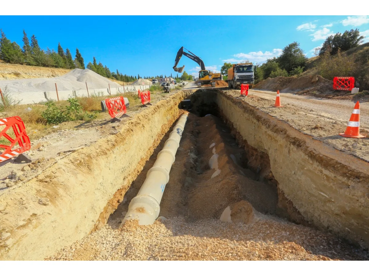 Açılan derin kazı kanalına beton borular döşeniyor, arkada iş makinesi ve kamyon var.