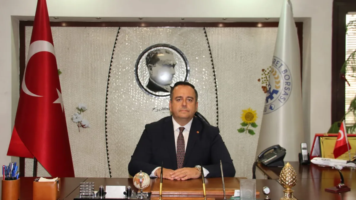 Makresinde oturan bir yetkili, arkasında Türk bayrağı, Denizli Ticaret Borsası bayrağı ve Atatürk portresiyle görülüyor.