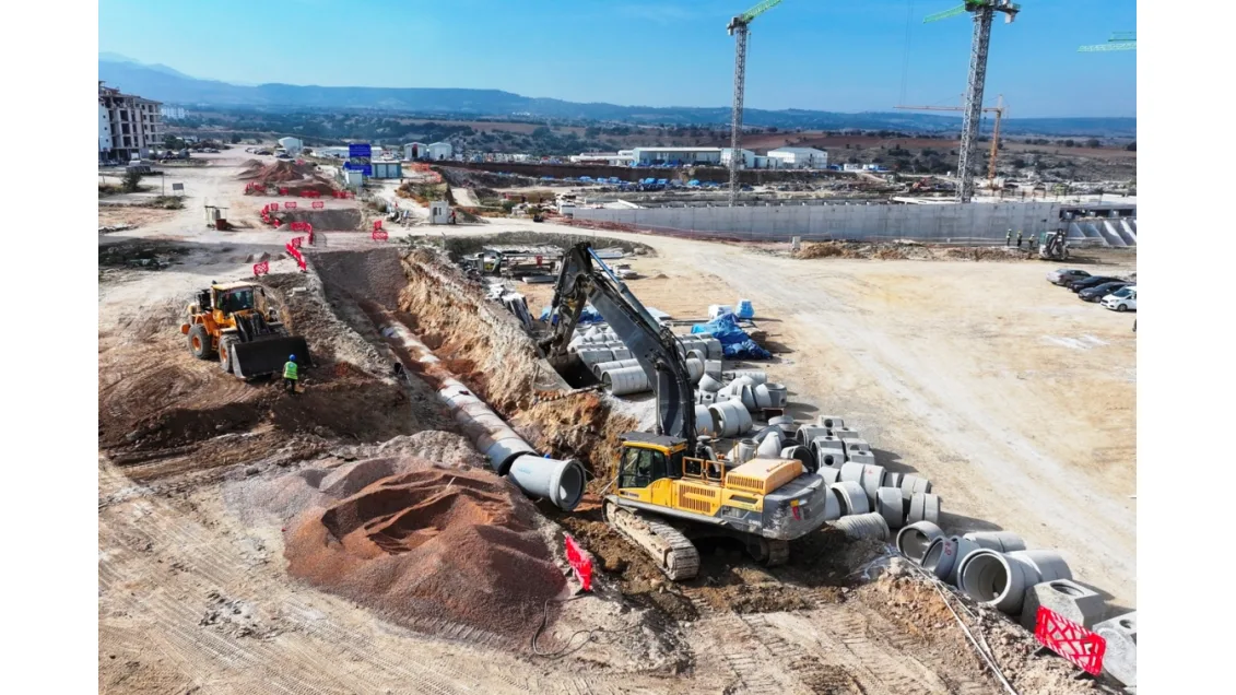 Şantiye alanında kazı yapan iş makineleri ve beton borular yer alıyor.