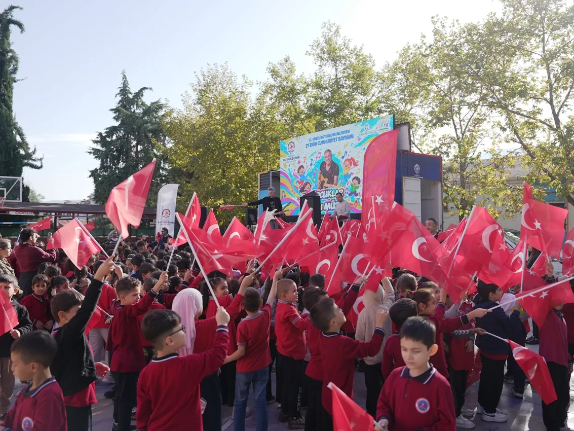 Okul bahçesinde ellerinde Türk bayrakları sallayan öğrenciler ve arka planda sahne.