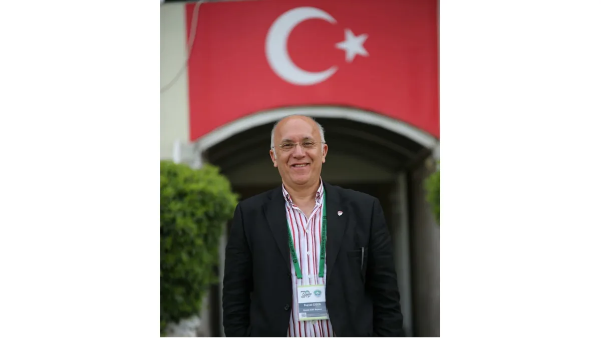 Arka planda Türk bayrağı asılı binanın önünde duran gözlüklü ve ceketli erkek.